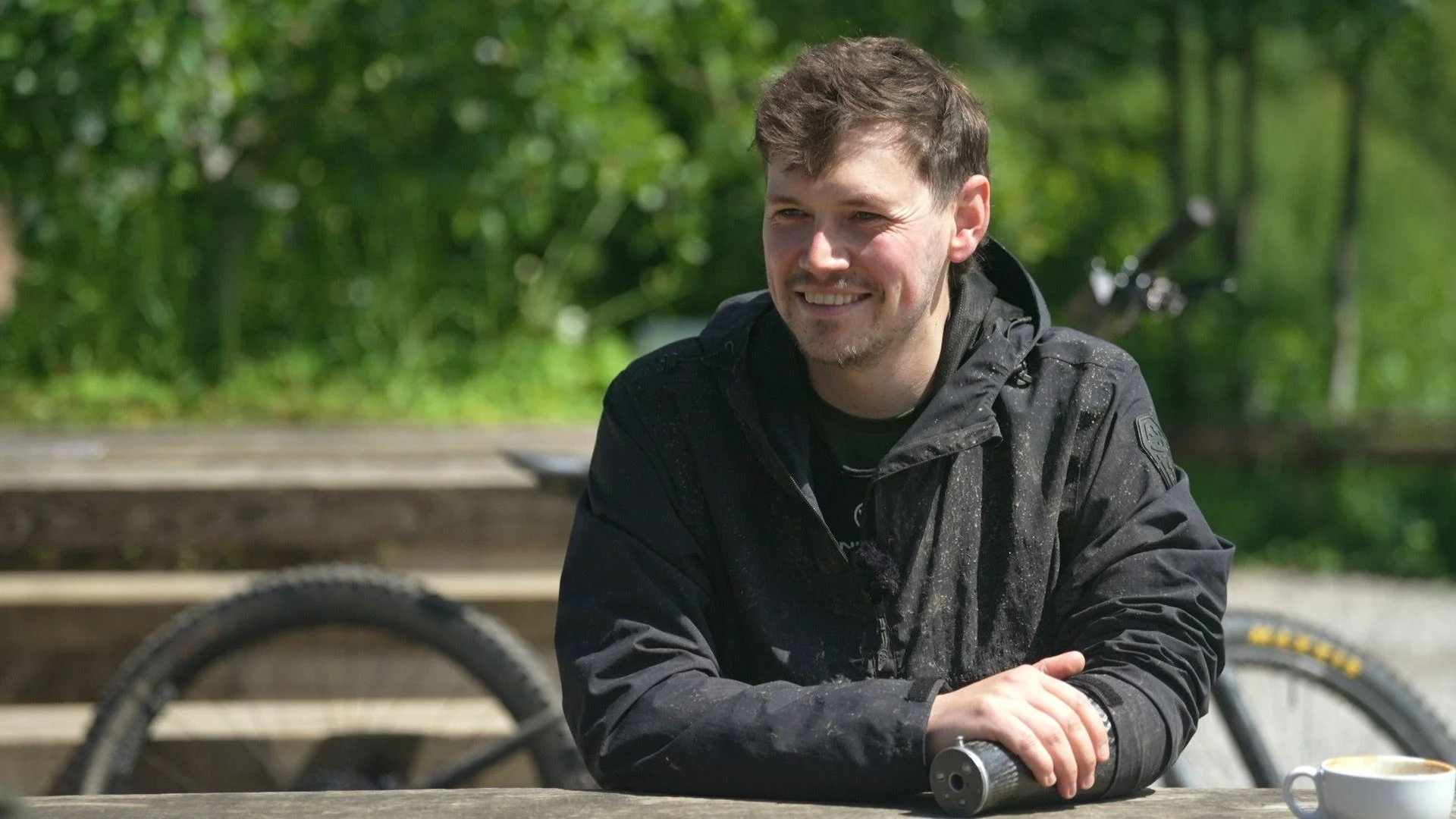 Morgan Jones wearing a black jacket and with a prosthetic left arm smiles at the camera, with a mountain bike in the background.