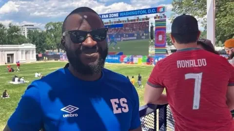 Ire Balogun wearing sunglasses and a blue shirt poses for a photo.