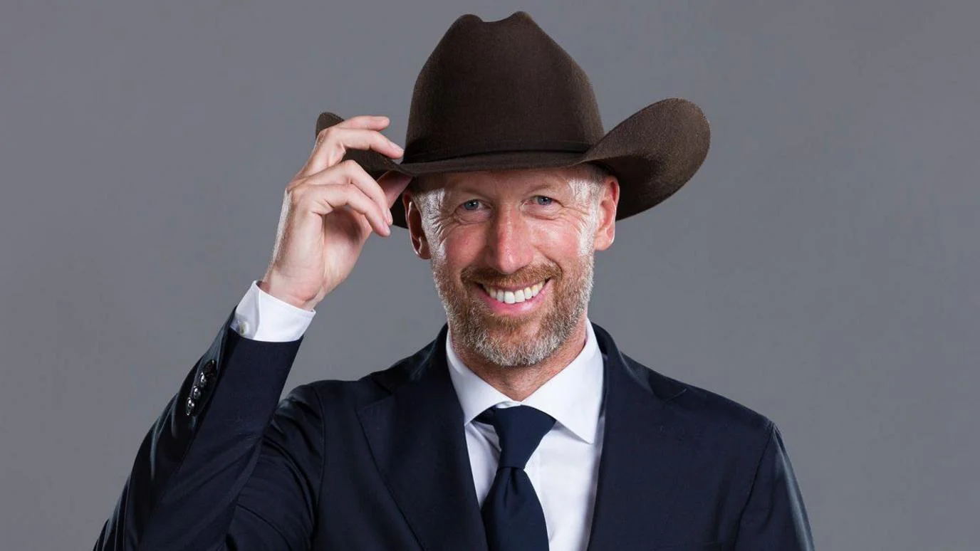 Graham Potter poses for the camera, wearing a suit while touching the brim of a cowboy hat